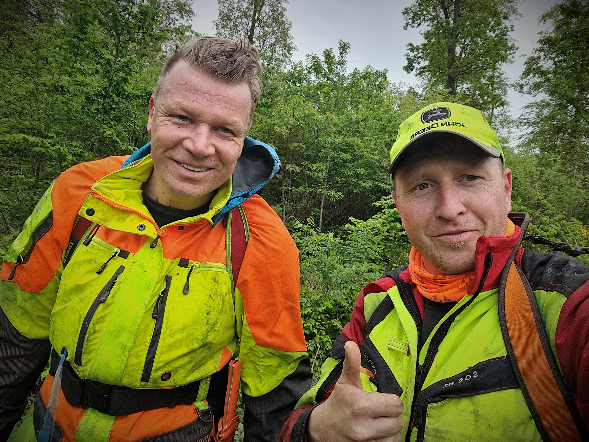 Zwei Männer in auffälliger Arbeitskleidung stehen in einer grünen Umgebung und lächeln in die Kamera.