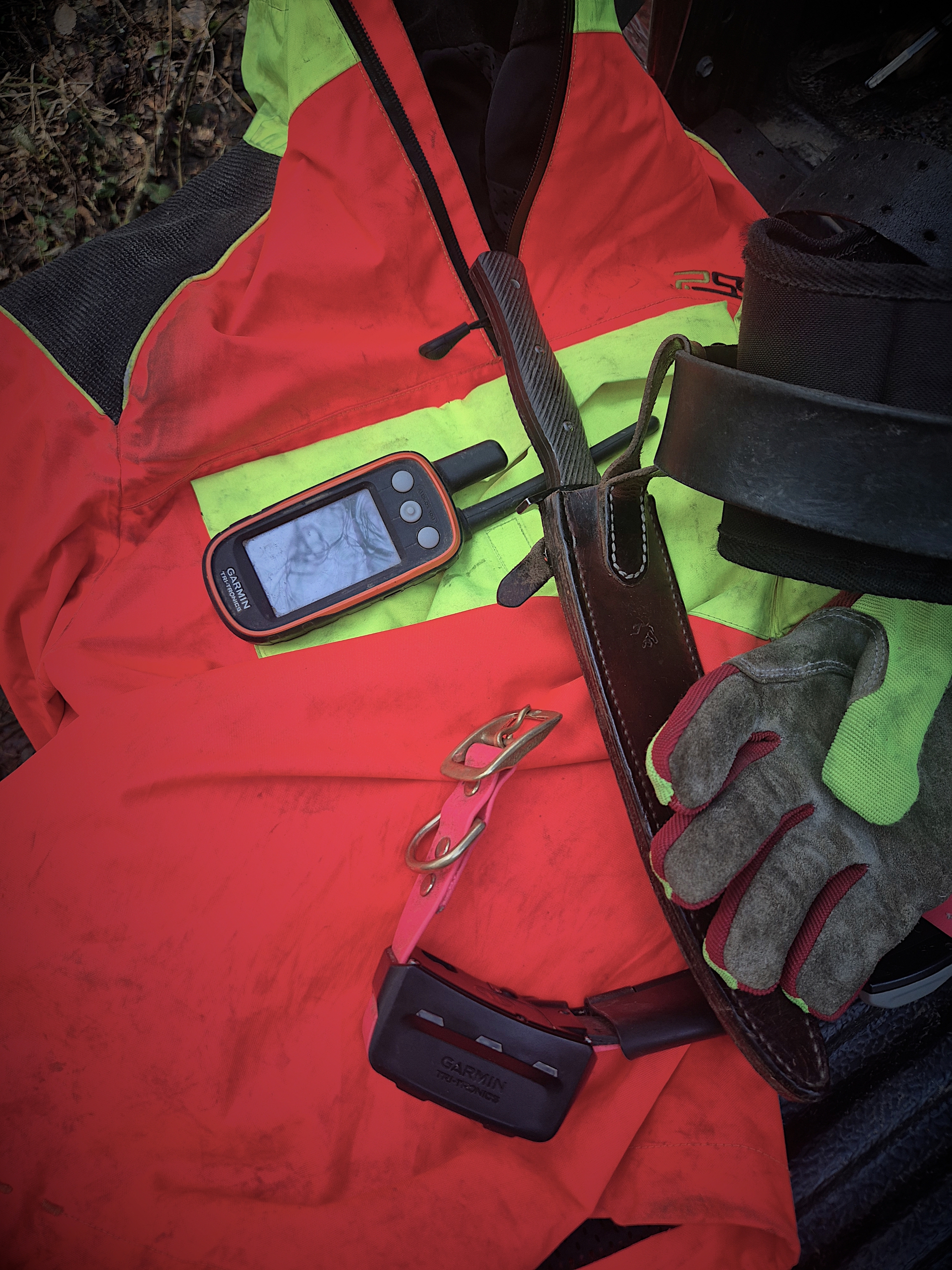 Rot-orange Jacke, GPS-Gerät, Handschuhe und ein Gürtel liegen auf einem Untergrund, dazu eine Waffe.