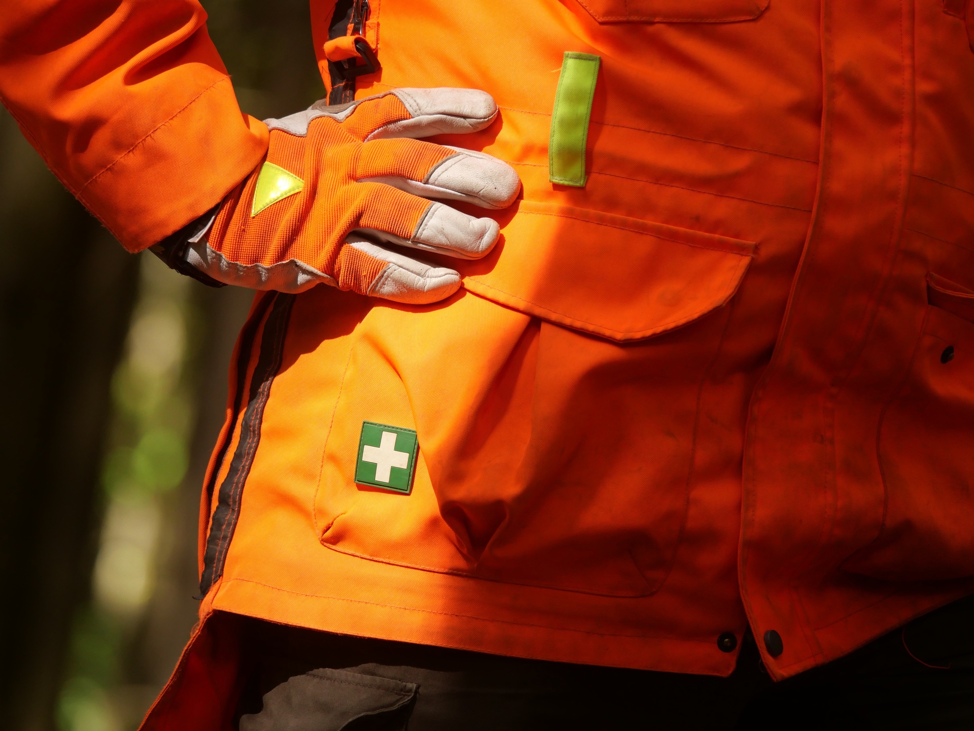 Ein Mann trägt einen orangefarbenen Arbeitsanzug mit grauen Handschuhen und einem Erste-Hilfe-Symbol auf der Tasche.